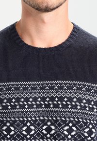 Mörkblå stickad tröja med rund halsringning, prydd med ett vitt geometriskt mönster över bröstet och textilier med struktur.