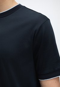 Gros plan d'une personne portant une chemise bleu marine foncé à manches courtes avec col côtelé et poignets ornés d'un liseré bleu clair.