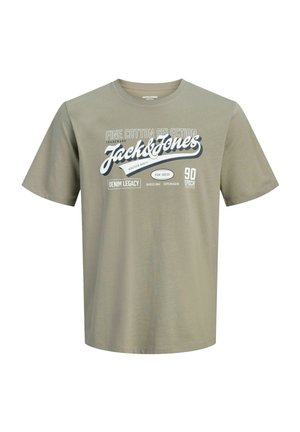 Teenage dreng med krøllet hår iført en grå Jack & Jones t-shirt, blå jeans og sorte sneakers står mod en ensfarvet baggrund.