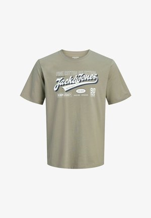 Adolescent aux cheveux bouclés portant un t-shirt gris Jack & Jones, un jean bleu et des baskets noires, debout devant un fond uni.