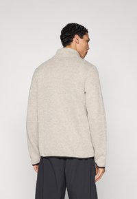 Μπεζ υφασμάτινο μπουφάν από φλις με ψηλό γιακά, ορατές ραφές και σκούρη επένδυση στο κάτω μέρος και στις μανσέτες.