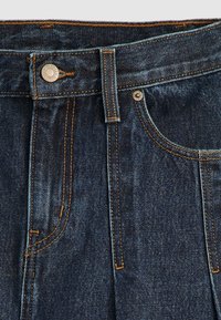 Gros plan de l'avant d'un jean en denim bleu foncé avec bouton métallique, passants de ceinture, poche avant et détails de couture orange.