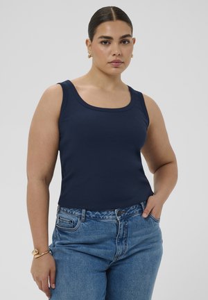 Woman with dark hair in a navy tank top and blue jeans, standing with one hand in pocket and wearing gold hoop earrings and bracelet.