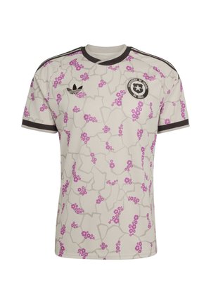 Camiseta de fútbol de color beige claro con racimos de flores rosas, líneas geométricas grises, cuello y ribetes de las mangas en negro, logo de Adidas y escudo de la Federación de Fútbol de Chile.
