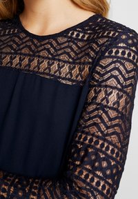 Marineblaues Kleid mit langen Spitzenärmeln und aufwendigen Mustern sowie einem Oberteil aus einfarbigem Stoff. Strukturierte Spitze verleiht dem oberen Teil Details.