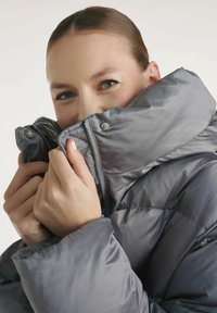 Kurtka puchowa w miękkim, szarym materiale z wysokim kołnierzem, z zamkiem błyskawicznym i zatrzaskami. Materiał ma gładką, błyszczącą fakturę z przeszyciami.