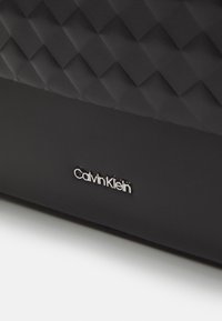 Borsa in ecopelle nera con texture trapuntata, caratterizzata da un logo Calvin Klein in argento. Superficie liscia con un design elegante e moderno.