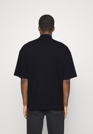 Ein Mann von hinten, der ein lockeres schwarzes T-Shirt und dunkle Jeans vor einem schlichten weißen Hintergrund trägt.