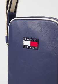 Námornícka modrá syntetická taška s hladkou textúrou, s logom v nášivke a červenými a bielymi akcentmi, a pruhovaným popruhom.
