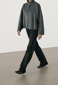 Veste en laine grise, courte, avec un col montant, une fermeture à boutons et des poches latérales, assortie à un pantalon noir et des chaussures plates.