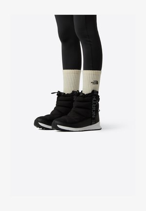 Cizme negre izolate cu partea superioară din material textil matlasat, talpă albă din cauciuc și logo negru pe lateral. Asortate cu șosete albe cu dungi și colanți negri.