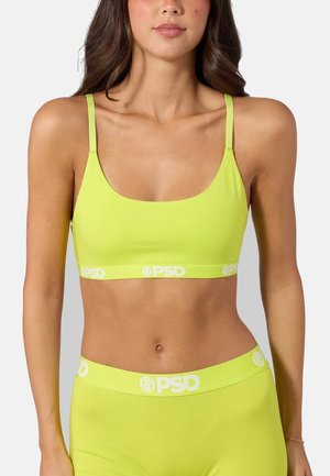 Kvinde iført limegrøn sports-bh og matchende high-waist underdele med hvide "PSD" logoer på elastikbånd mod en ensfarvet baggrund.