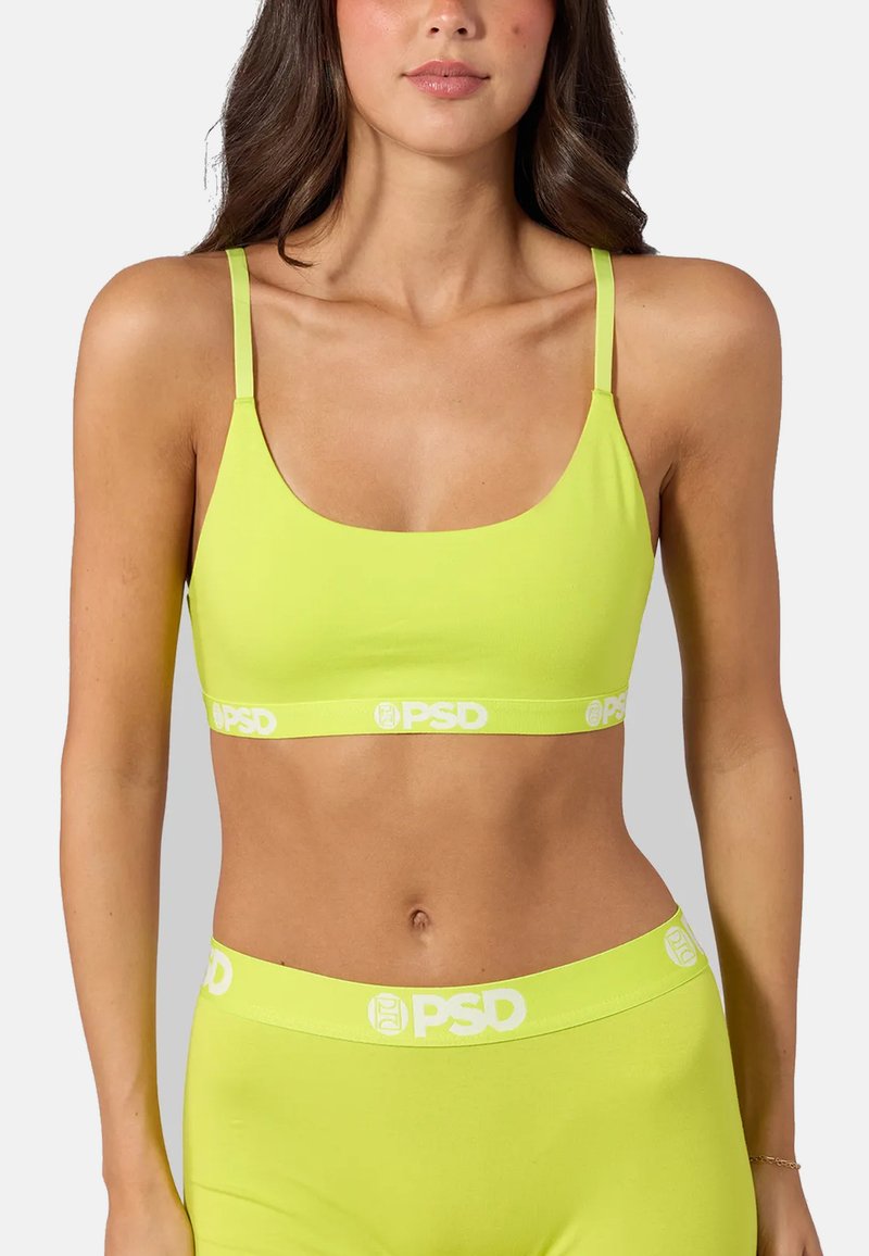 Femme portant un soutien-gorge de sport vert citron vert et un bas taille haute assorti avec des logos blancs "PSD" sur les bandes élastiques sur fond uni.