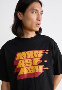 Black cotton t-shirt featuring bold, stylized graphic in yellow and red that reads "MAG ASH" with fluid, dynamic lettering.