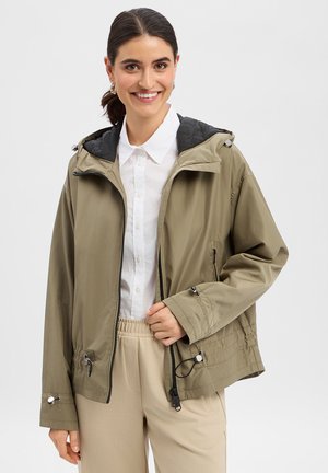 Lächelnde Frau, die eine khakifarbene Kapuzenjacke über einem weißen Hemd mit Knöpfen und beigefarbenen Hosen trägt, vor einem schlichten hellen Hintergrund posierend.