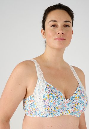 Femme portant un soutien-gorge à motif floral avec des bretelles en dentelle, posant devant un fond gris clair uni.