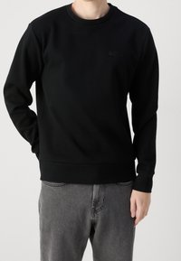 Osoba w czarnym długim rękawie sweatshirt BOSS i szarych dżinsach, stojąca z jedną ręką częściowo w kieszeni na jednolitym tle.