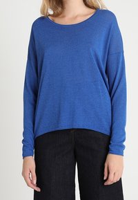 Top blu lungo con maniche a sbuffo realizzato in un tessuto morbido e leggero. Presenta una scollatura rotonda e una vestibilità comoda. Abbinato a pantaloni in denim scuro.