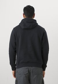 Sweatshirt preto com capuz, em mistura de algodão, com punhos e bainha canelados, design liso, usado sobre calças cargo cinzentas, mostrado por trás.