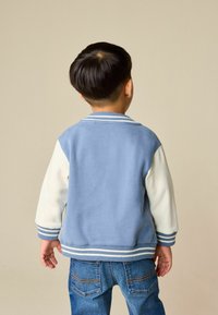 Niebiesko-biała kurtka w stylu varsity o miękkiej fakturze, z prążkowanymi mankietami i paskowanym dołem, zestawiona z niebieskimi dżinsami. Widok dziecka od tyłu.