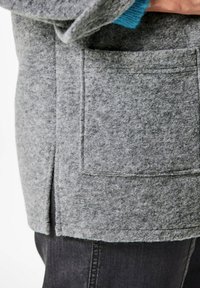 Grauer Wollmantel mit sichtbarer Tasche, seitlichem Schlitz und strukturierter Oberfläche. Eine blaue Strickschicht ist ebenfalls unter dem Ärmel sichtbar.
