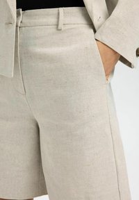 Pantaloni corti in lino beige con un design sartoriale, dotati di tasche laterali e una texture liscia. Adatti per l'abbigliamento estivo.