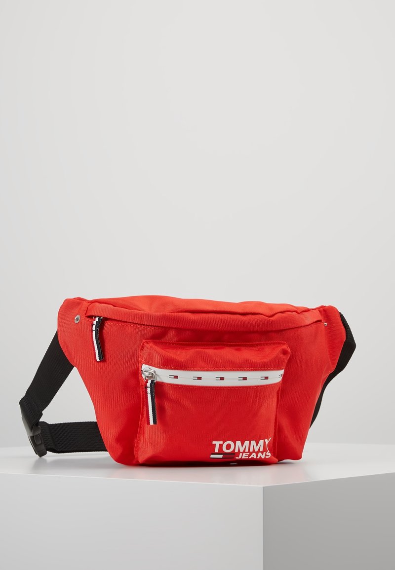 Rode Tommy Jeans heuptas met een voorvak met rits, zwarte verstelbare band en wit logo op een witte standaard.