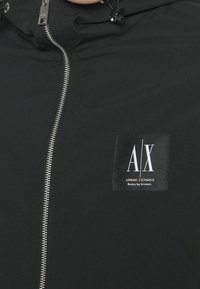 Svart jacka med full dragkedja, justerbar dragskohuva och en fyrkantig logotyp med texten "A/X" på vänster bröst. Slät tygtextur.