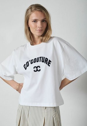 Blondynka ubrana w biały, oversize'owy T-shirt z napisem "CO'COUTURE" oraz beżowe spodnie w paski, stoi z rękami na biodrach, neutralne tło.