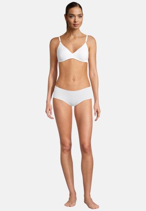 Frau mit heller Haut und dunklem Haar, die einen weißen BH und Hipster-Unterwäsche trägt, barfuß vor einem schlichten hellen Hintergrund steht.