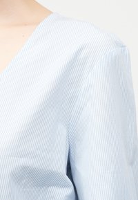 Κοντινό πλάνο σε μανίκι και ώμο πουκάμισου με κάθετες ρίγες σε ανοιχτό γαλάζιο και άσπρο, σε φόντο από γυμνό δέρμα.