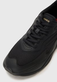 Black sneaker featuring mesh and synthetic materials, round toe, thick sole, and logo detail at the tongue in gold. Textured upper with laces.