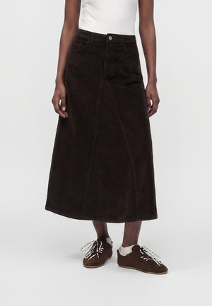 Personne portant une jupe midi en velours côtelé marron foncé, de forme trapèze, des chaussettes blanches, des chaussures marron à lacets et un top blanc sans manches sur un fond uni.
