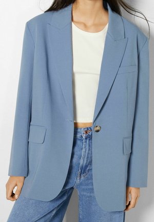 Woman wearing light blue single-button blazer over white ribbed crop top and high-waisted blue jeans, standing against a plain background.