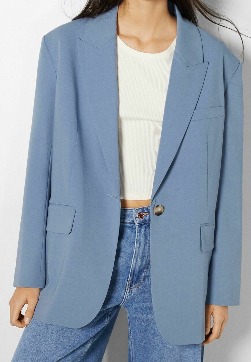 Femme portant un blazer bleu clair à bouton unique sur un crop top blanc côtelé et un jean bleu taille haute, debout devant un fond uni.