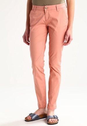 Pantalon slim rose saumon porté par une personne debout, associé à des sandales bleu marine avec détail de boucle sur fond blanc.