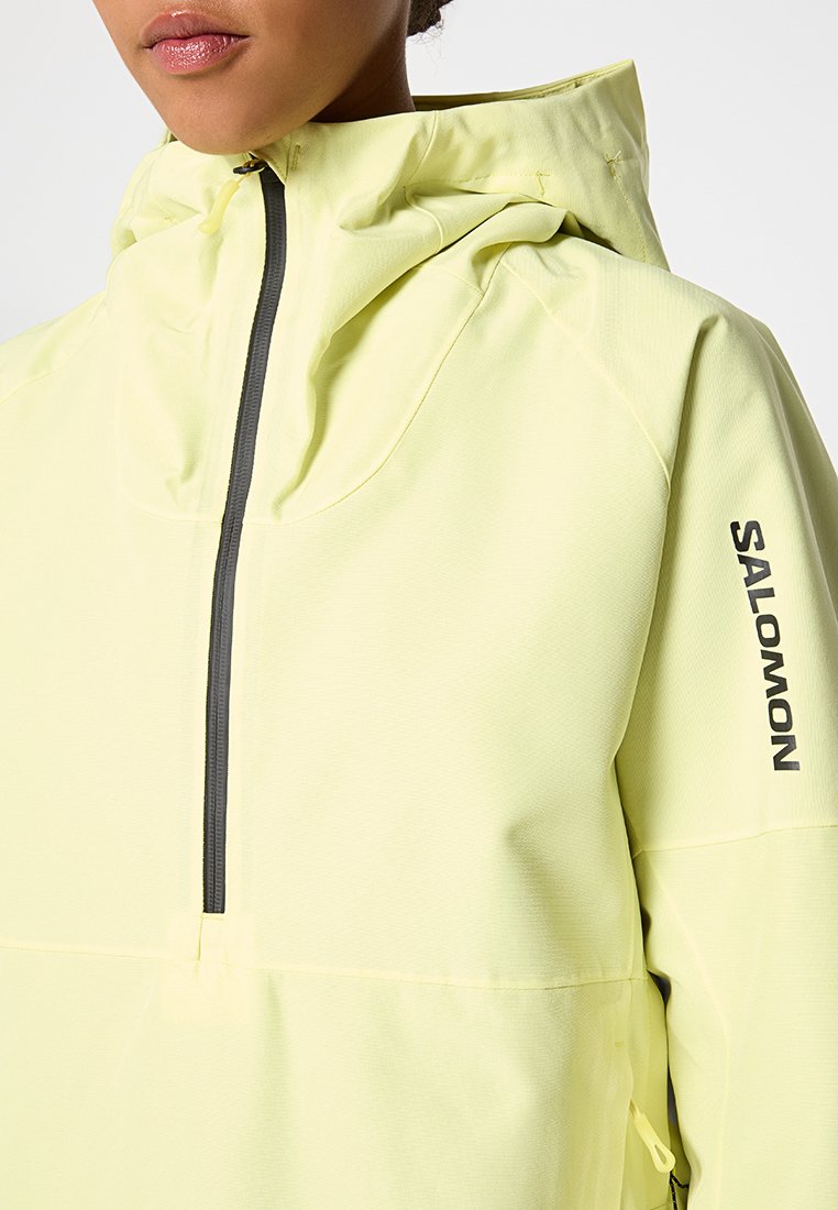 Chaqueta de pullover amarilla clara con un alto cuello, media cremallera y logotipo negro en la manga. La tela tiene un acabado suave y texturizado.