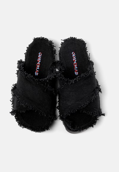 Svarta tygslip-ons med fransiga korsade remmar, texturerad innersula och en solid svart sula. Branderlogotyp tryckt i kontrasterande färger.