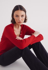 Rode longsleeve top met een ribbeltextuur en een drieknoopsluiting, gecombineerd met strakke zwarte jeans. Model zit met gekruiste benen, armen op de knieën rustend.