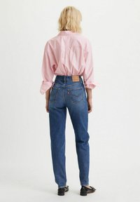 Camisa de botones a rayas en rosa claro, jeans de tiro alto azules con un parche de logo en la parte trasera, y bailarinas negras, vista desde la parte de atrás.