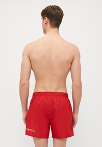 Calções de banho vermelhos com cintura elástica e detalhe de logotipo branco. Textura suave, design até aos joelhos, adequado para vestuário de praia casual.