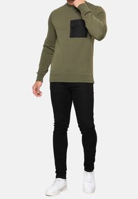 Olivově zelená mikina s černou náprsní kapsou, kulatým výstřihem, přiléhavým střihem, doplněná černými úzkými džínami a bílými teniskami.