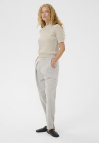 Femeie cu păr creț, îmbrăcată cu un pulover bej cu mânecă scurtă, pantaloni gri deschis, cu mâinile în buzunare și pantofi negri fără șireturi, stând de profil.