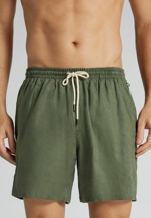 Persona sin camiseta llevando pantalones cortos verde oliva con cordón y cintura elástica, manos relajadas a los lados, mostrada de la cintura hasta mitad del muslo.