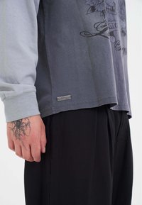 Grijs en lichtblauw langarm shirt met een grafische print, met een metalen label aan de onderkant en een zwarte achtergrond.