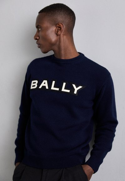 Navy blue knitted sweater featuring a white "BALLY" logo on a black patch. Ribbed neckline and cuffs. Model depicted in profile view.