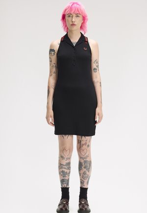 Persona con cabello rosa y múltiples tatuajes lleva un vestido polo negro sin mangas con botones y zapatos negros sobre un fondo liso.