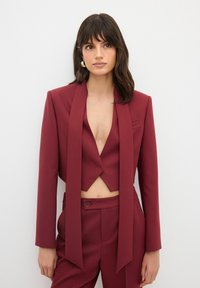 Burgundy kostymset med en kort blazer med djup V-ringning och lång, draperad scarf-detalj, samt högmidjade byxor med knäppning.