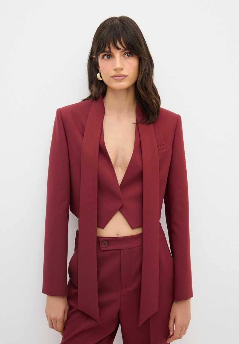 Burgundy kostymset med en kort blazer med djup V-ringning och lång, draperad scarf-detalj, samt högmidjade byxor med knäppning.