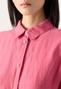 Camicia in lino rosa con colletto casual, caratterizzata da una finitura testurizzata e chiusura frontale con bottoni. I dettagli includono bottoni morbidi e una vestibilità rilassata.